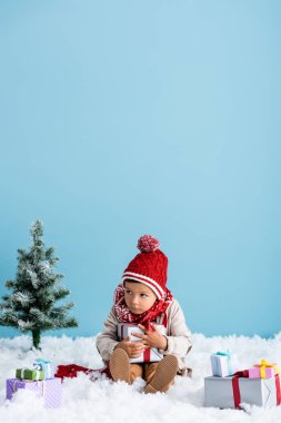 Şapkalı ve kışlıklı çocuk karda oturuyor ve mavi üzerine izole edilmiş yılbaşı ağaçlarının yanında hediye kutusu tutuyor.