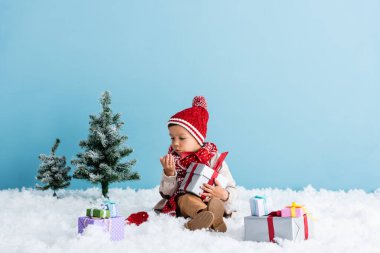 Şapkalı ve kışlıklı bir çocuk karda oturuyor ve hediyesini tutuyor. Elinde esinti var. Mavi renkte.