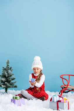 Şapkalı ve kışlıklı bir çocuk karda oturuyor ve mavi kızağın yanında hediyesini tutuyor.