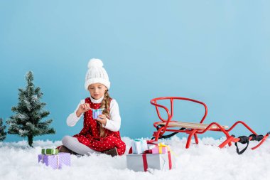 child in hat and winter outfit sitting on snow and holding present near sleigh isolated on blue clipart