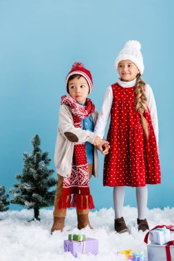 Kız kardeşiyle el ele tutuşan örülü şapkalı çocuk mavi üzerine izole edilmiş kar üzerinde hediyeler veriyor.