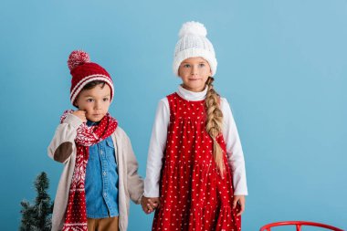 kız kardeşiyle el ele tutuşurken, mavi şapkalı ve eşarplı çocuk yanağa dokunuyor.