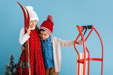 Kışlık kıyafetli kız elinde kayak ve kucaklaşan erkek kardeş, maviliğin yanında tek başına duruyor. 