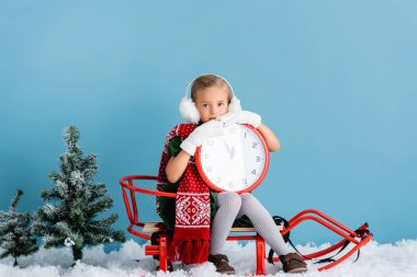 girl in winter earmuffs and scarf sitting in sleigh and holding clock near pines on blue clipart