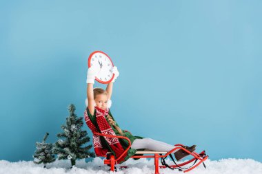 girl in winter earmuffs and scarf sitting in sleigh and holding clock above head on blue clipart