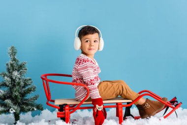 boy in winter earmuffs and sweater sitting in sleigh on snow near pine on blue clipart