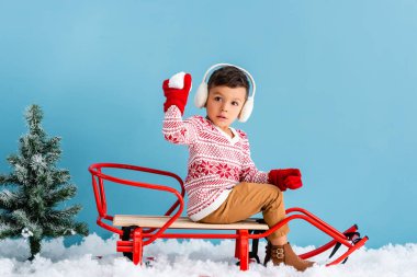 boy in winter earmuffs and sweater sitting in sleigh and holding snowball on blue clipart