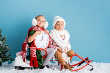 kids in winter outfit and scarfs sitting on sleigh near clock, pine and ice skates on blue clipart