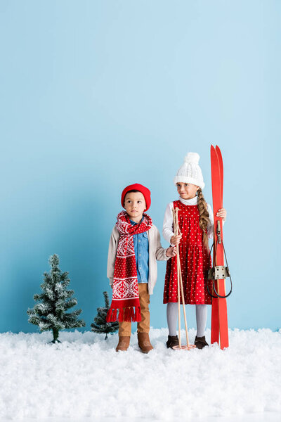girl holding ski poles and skis while standing on snow near brother in winter outfit on blue