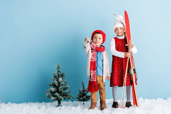 boy in hat and scarf pointing with finger near sister holding ski poles and skis while standing on blue