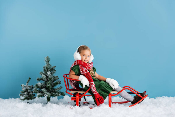 girl in winter earmuffs and scarf sitting in sleigh on snow near pines on blue