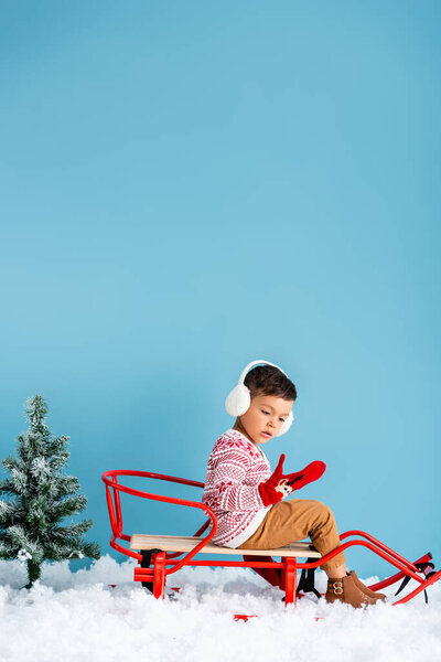 boy in winter earmuffs and sweater sitting in sleigh on snow near pine on blue