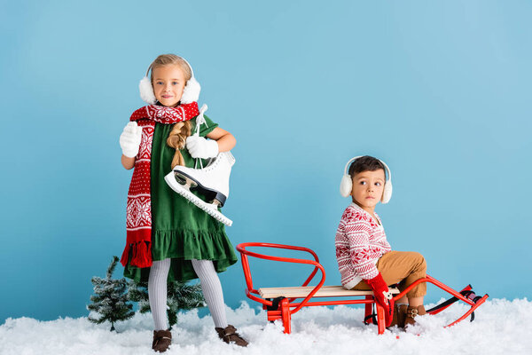 girl in winter earmuffs and scarf standing with ice skates near brother in sleigh on blue