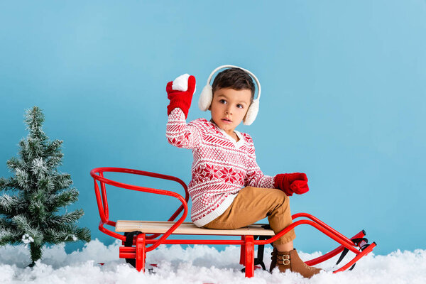 boy in winter earmuffs and sweater sitting in sleigh and holding snowball on blue