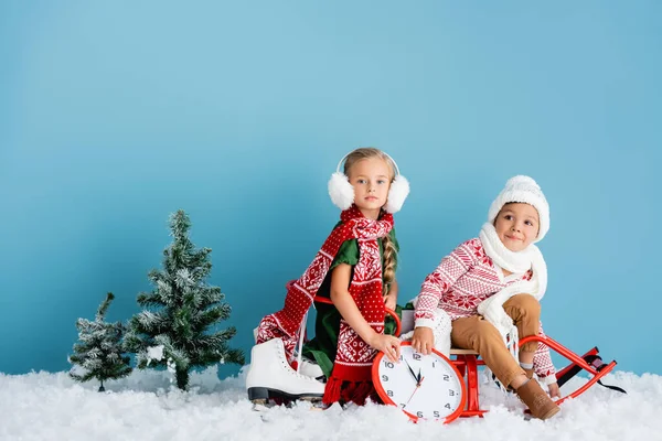 穿着围巾和冬装的孩子们坐在雪橇上接近时钟 松木和冰鞋穿着蓝色 — 图库照片