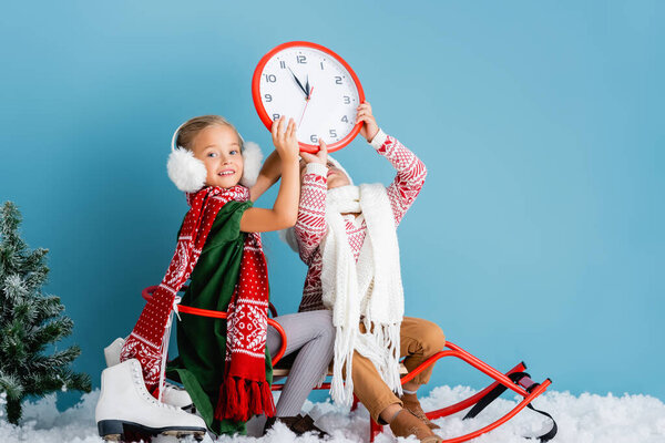 kids in winter outfit sitting on sleigh and holding clock near pine and ice skates on blue