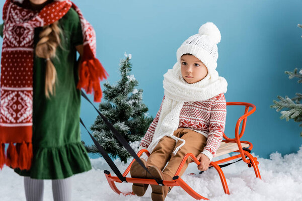 selective focus of boy in winter outfit sitting on sleigh near sister on blue