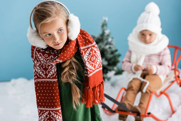 selective focus of girl in winter outfit giving a ride to boy on sleigh on blue
