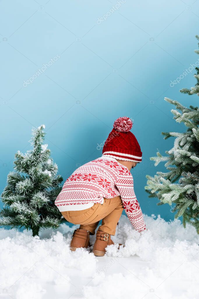 boy in winter outfit sitting near white snow near christmas trees on blue