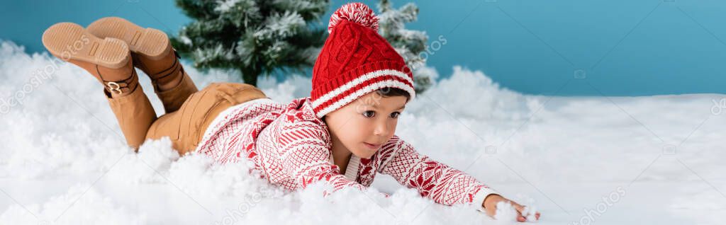 panoramic concept of boy in winter outfit lying on white snow near christmas trees on blue
