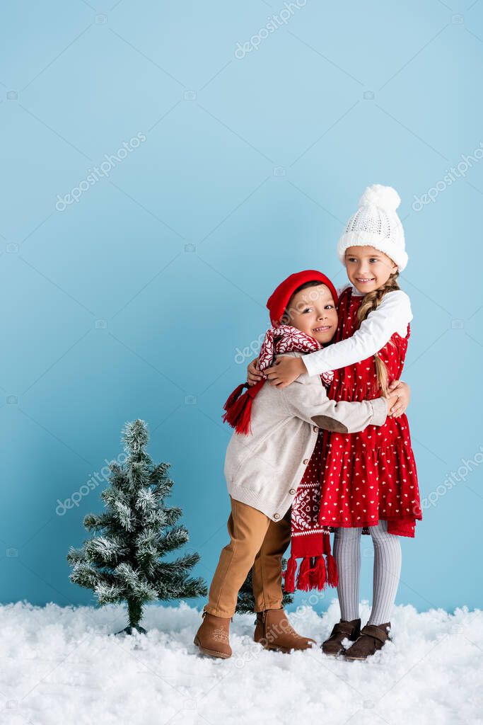 children in winter outfit hugging near christmas tree on blue