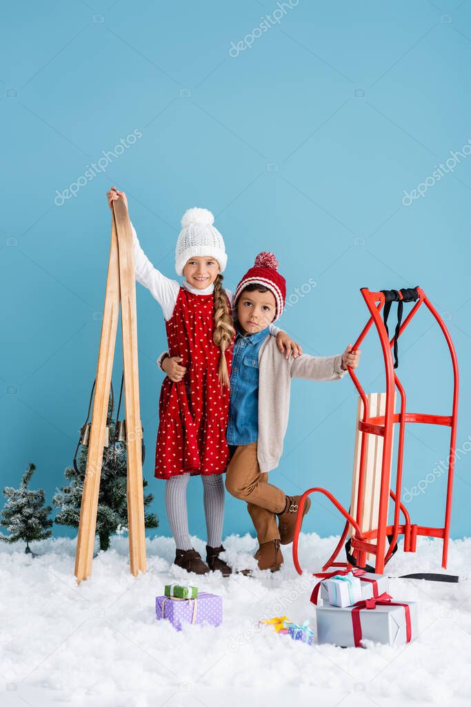 girl in winter outfit holding skis and hugging brother standing near sleight and presents on snow isolated on blue 