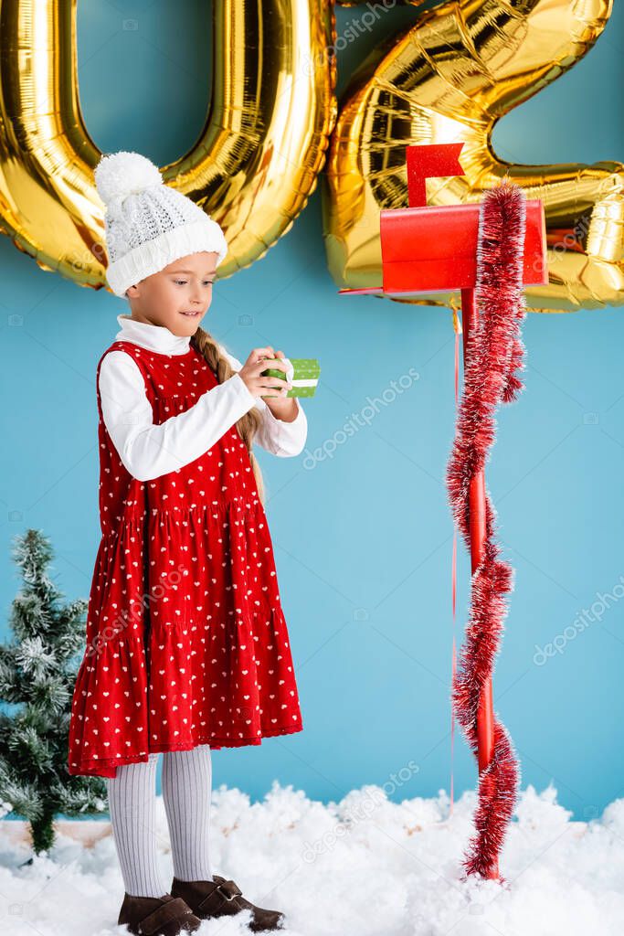 girl in hat holding present while standing near mailbox and balloons on blue