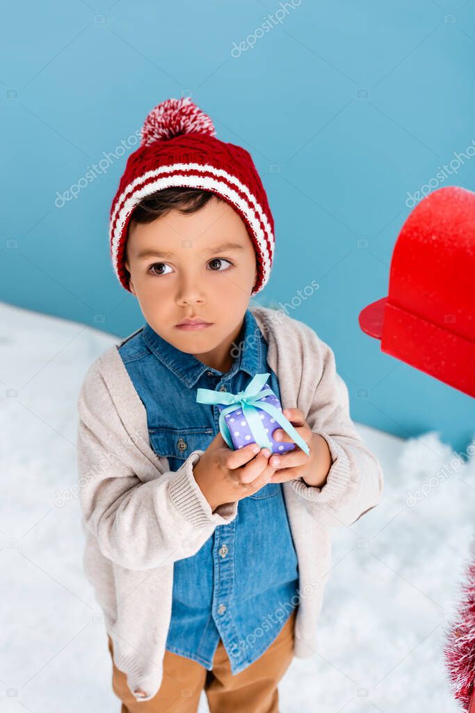 boy in hat and winter outfit holding present near red mailbox on blue 