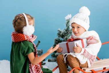 Örgü şapkalı çocuk, mavi kulaklıklarla el kol hareketi yapan bir kızın yanında otururken hediyesini tutuyor.