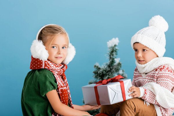 selective focus of girl in winter earmuffs near boy in knitted hat holding present on blue