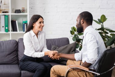 joyful patient shaking hands with african american psychologist while sitting on sofa clipart