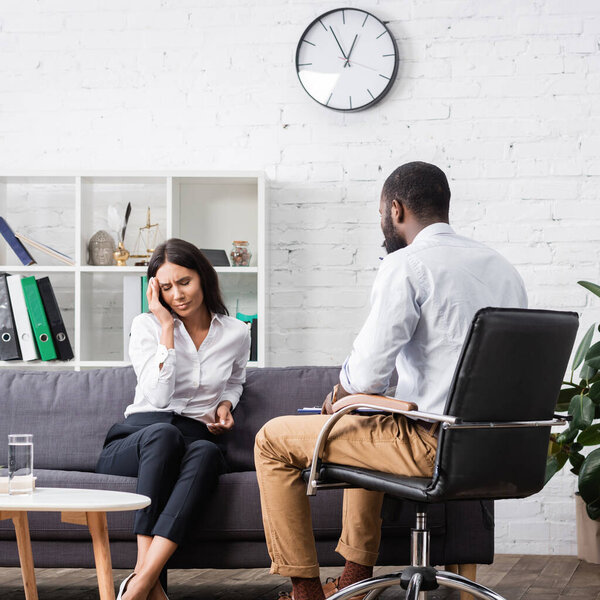 sad woman touching head while sitting on sofa with closed eyes near african american psychologist