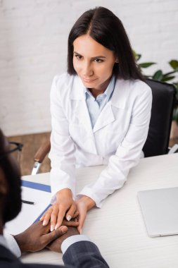 Esmer doktorun, iş yerinde otururken Afrikalı Amerikalı bir hastanın ellerine dokunmasının seçici odağı.