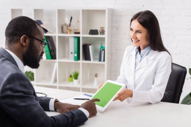 Seçici odak noktası, Afrikalı Amerikalı bir adamın dijital tableti işaret etmesi ve yeşil ekranlı esmer bir doktorun yanında olması.