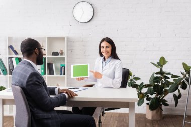 Esmer doktor kameraya bakarken Afro-Amerikan hasta yakınındaki yeşil ekranlı dijital tableti işaret ediyor.