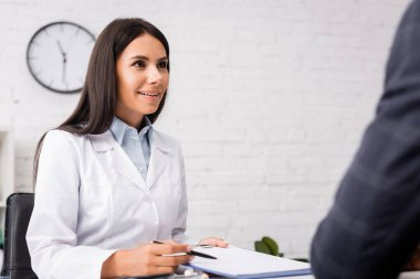 selective focus of brunette doctor holding clipboard and pen near african american patient clipart