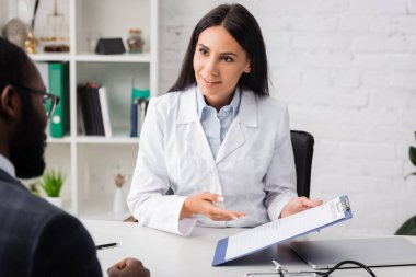selective focus of brunette doctor pointing with hand at clipboard with insurance form near african american man clipart