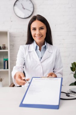 Esmer doktor elinde sigorta talep formuyla elinde kalem ve pano tutarken kameraya bakıyor.