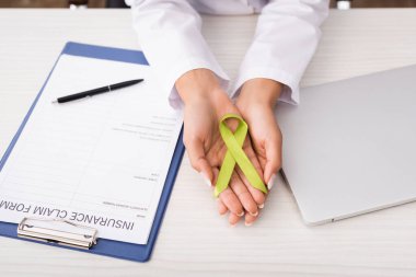 cropped view of psychologist holding green awareness ribbon near insurance claim form and laptop, mental health concept clipart
