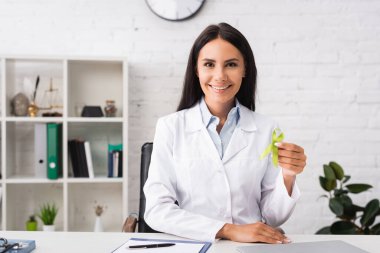 joyful doctor looking at camera while presenting green awareness ribbon, mental health concept clipart