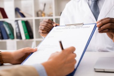 cropped view of patient signing insurance claim form near african american doctor clipart