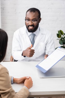 Gözlüklü Afrikalı Amerikalı doktorun seçici odak noktası elinde sigorta talep formu tutarken başparmağını kaldırması. 