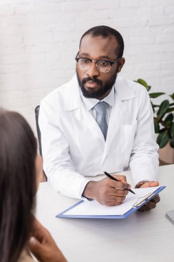 Gözlüklü Afrikalı Amerikalı doktor hasta bir hastanın boğazına dokunurken elinde not panosu ve kalem tutuyor.