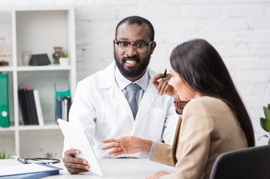 Afro-Amerikan doktor elinde dijital tabletle kameraya bakıyor ve esmer kadını işaret ediyor.