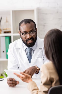 Gözlüklü Afrikalı Amerikalı doktor hasta ile konuşurken dijital tableti işaret ediyor.