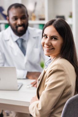 Afro-Amerikan doktorun yanındaki kameraya bakan neşeli esmer kadının seçici odağı.