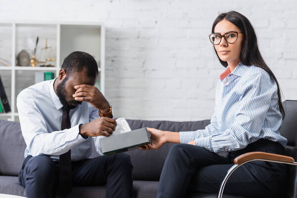 Брюнетка психолог в очках давая бумажные салфетки плачущий африканский американец закрывая глаза рукой