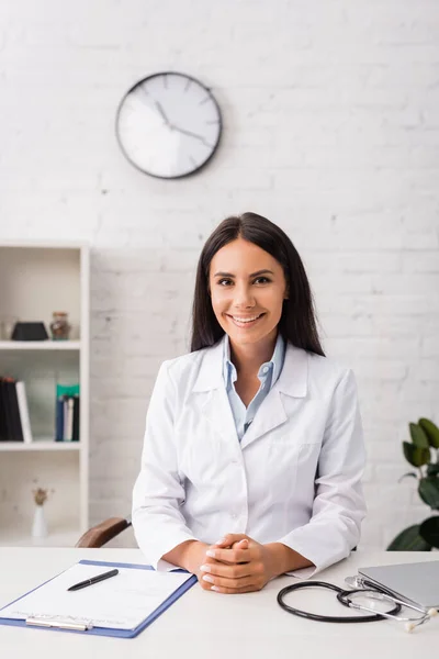 joyful brunette doctor looking at camera while sitting at workplace near clipboard with insurance form