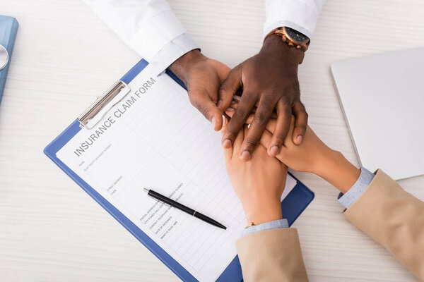 top view of clipboard with insurance claim form and african american doctor touching hands of patient