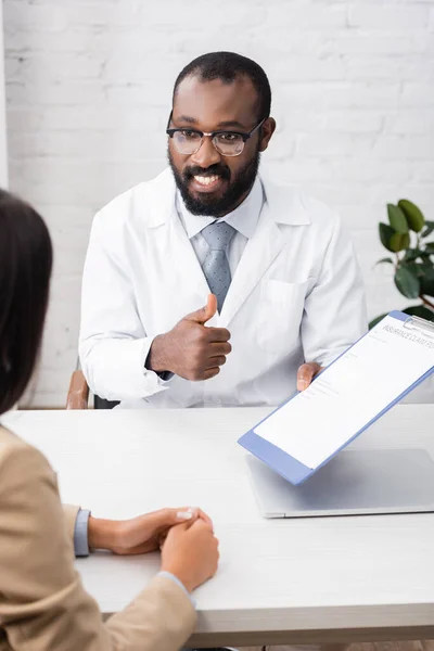 保険金請求書を持ちながら親指を立てる眼鏡のアフリカ系アメリカ人医師の選択的焦点 — ストック写真